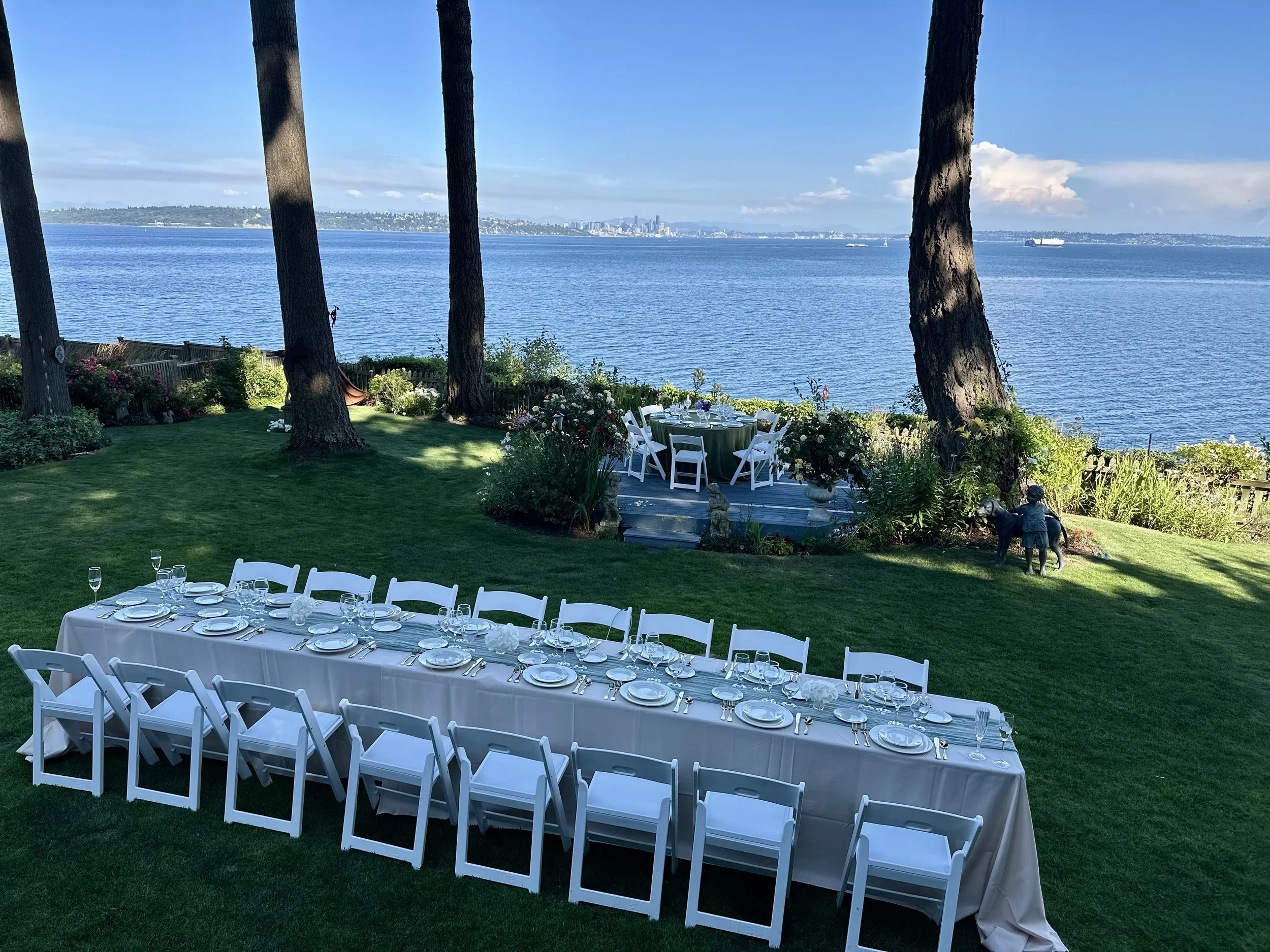 Long dining table set with white tablecloth, plates, glassware, and cutlery, arranged outdoors on well-manicured grass, with a view of the water and city skyline in the background, surrounded by trees and flowers.