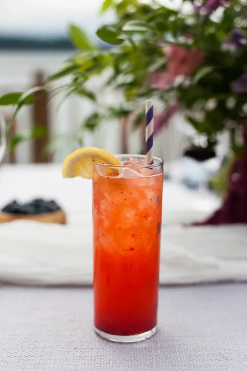 A tall glass of bright red fruit drink with ice, garnished with a lemon wedge and a striped straw, on a white table with blurred flowers and outdoor background.