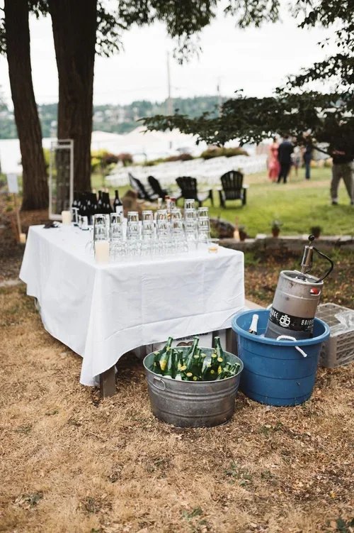 Outdoor gathering setup with a table covered with a white cloth, glassware, and bottles, surrounded by trees and people in the background, with buckets of bottles and a keg nearby.