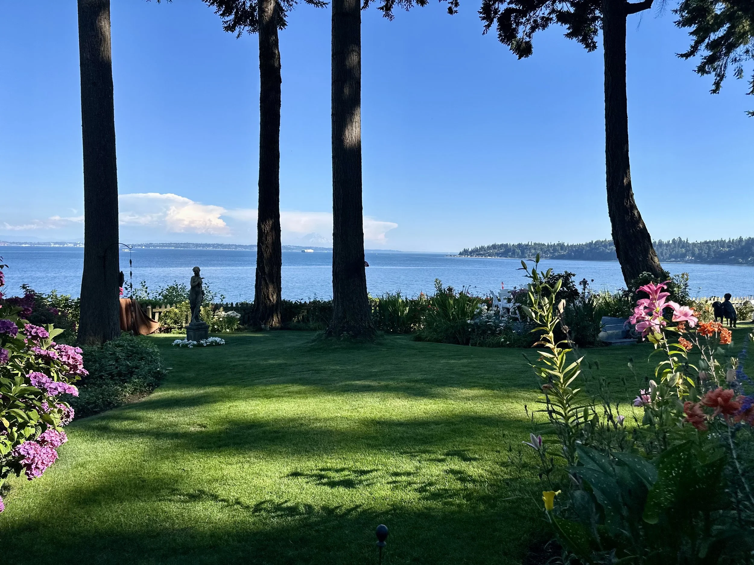 A peaceful garden with lush green grass, colorful flowers, and tall trees overlooking a body of water, with a distant landmass and a partly cloudy sky in the background.