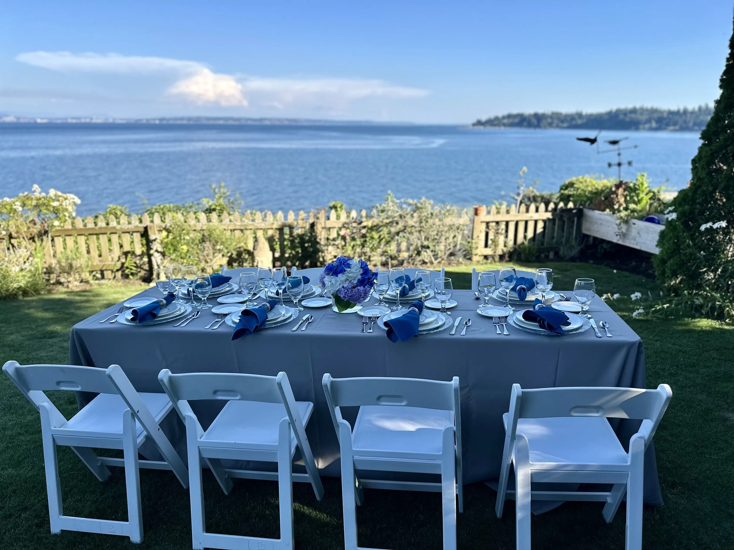 Wedding reception setup with a long table, white chairs, and blue napkins, overlooking a body of water with a fence and greenery in the background.