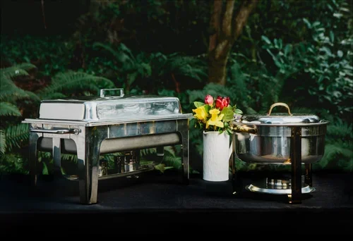 Buffet setup with two silver chafing dishes and a white vase with yellow and pink flowers, set on a black table with a forest background.