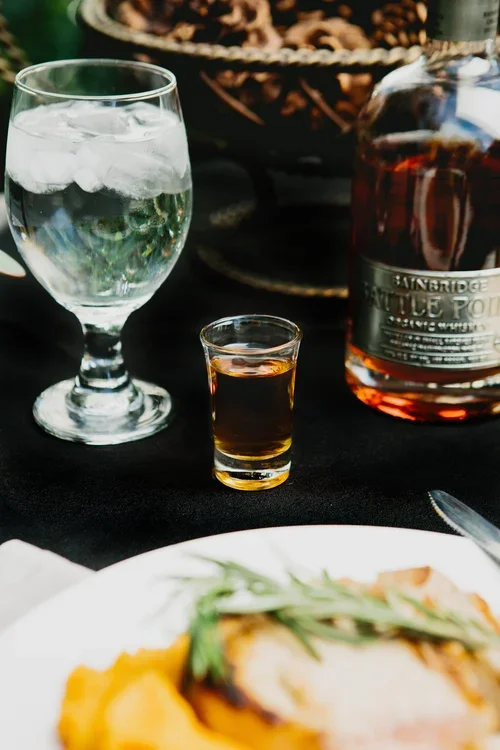 A glass of water with ice, a shot glass of dark liquor, and a bottle of bourbon on a table. Part of a plate with food is visible at the bottom.