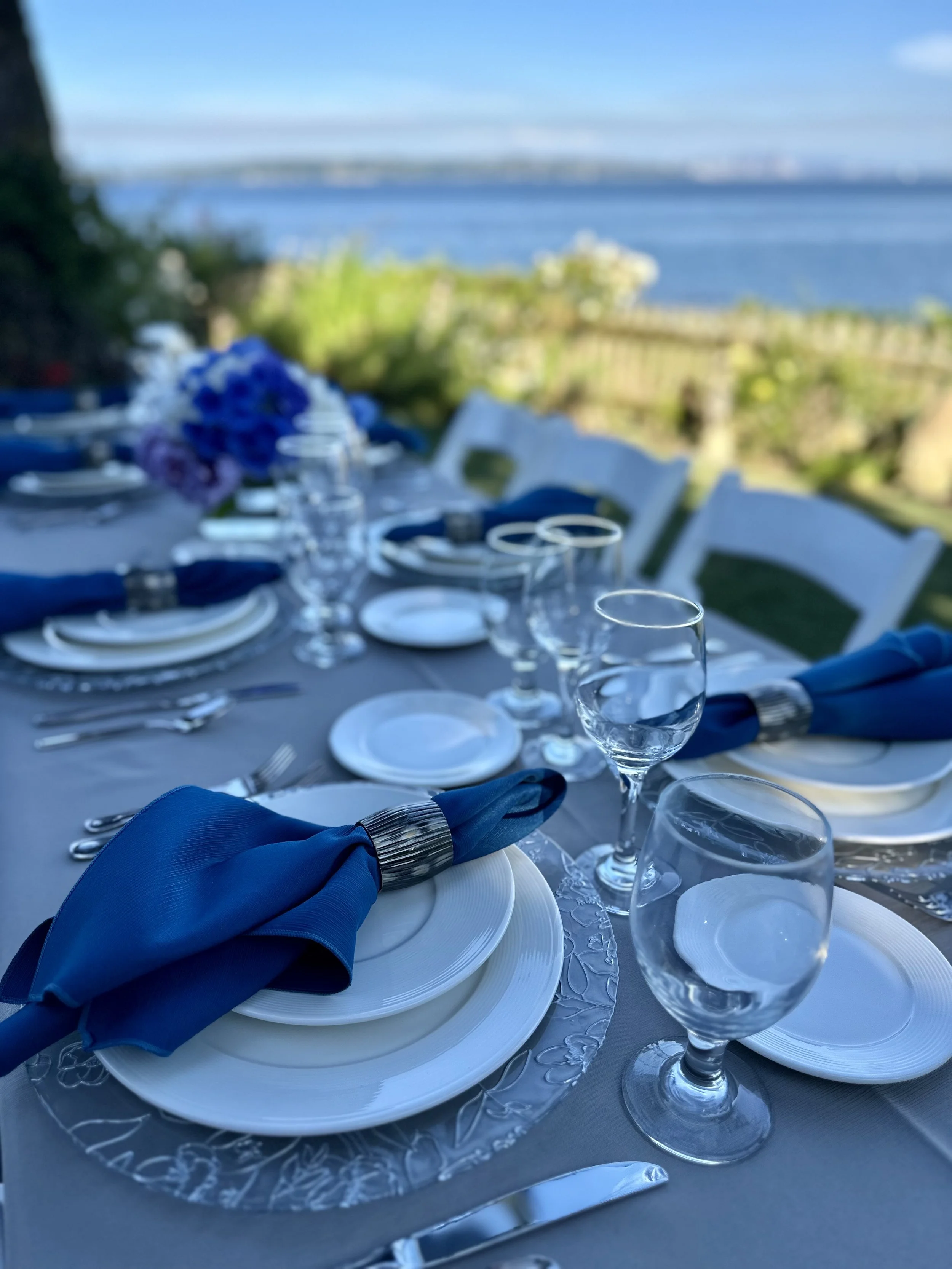 A formal outdoor dining table set with white plates, silverware, wine glasses, and blue cloth napkins, with a view of a body of water and a landscape in the background.