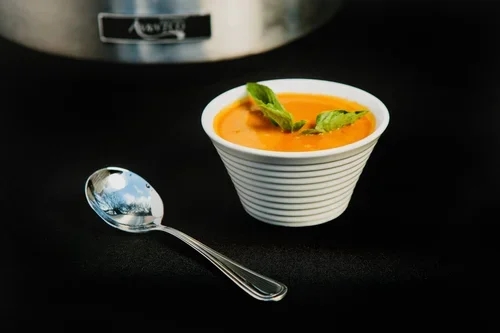 A bowl of creamy orange-colored soup garnished with fresh basil leaves, placed on a black surface with a silver spoon nearby.
