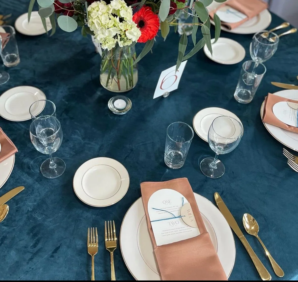 Elegant table setting with gold flatware, pink napkins, white plates, clear glassware, and a floral centerpiece with white hydrangeas, green foliage, and a red flower, on a dark blue tablecloth.