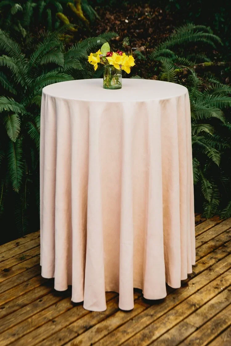 A round table covered with a white tablecloth, with a small vase of yellow and pink flowers, on a wooden floor with green plants in the background.