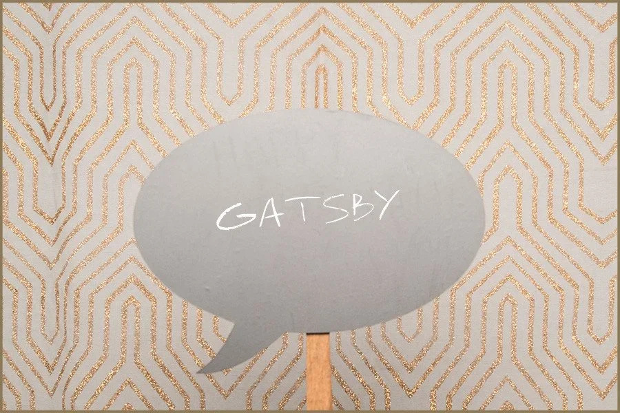 A decorative sign shaped like a speech bubble with the word 'Gatsby' written inside, set against a gold and white geometric patterned background.