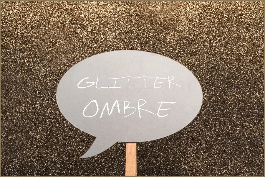 A speech bubble sign with the words 'GLITTER OMBRE' written on it, set against a glittery gold background.