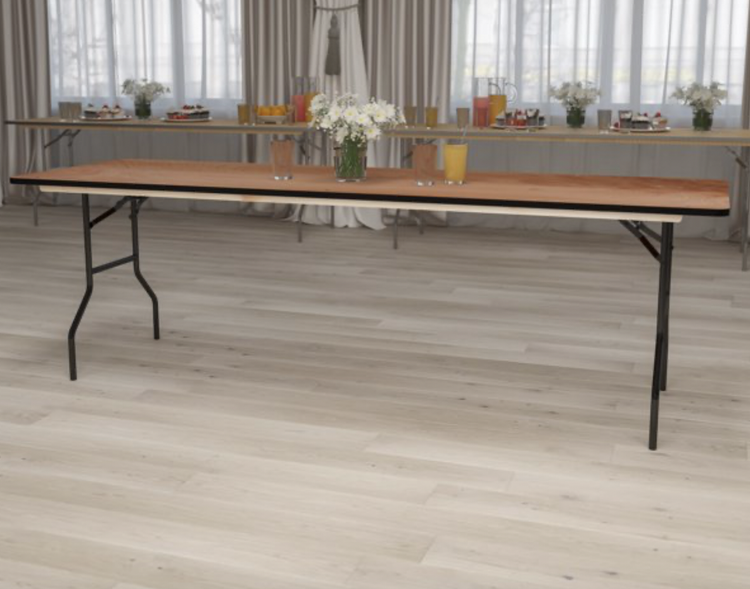 A long dining table with a wooden top and metal legs, set with flower arrangements, cups, and plates, in a room with light-colored wooden floors and large windows with curtains.