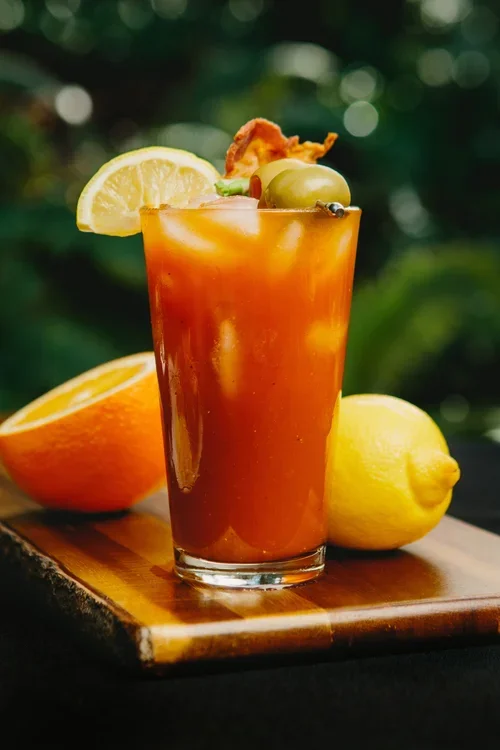 A glass of Bloody Mary garnished with lemon, green olives, and a crispy bacon strip, set on a wooden surface with lemon and orange halves in the background.