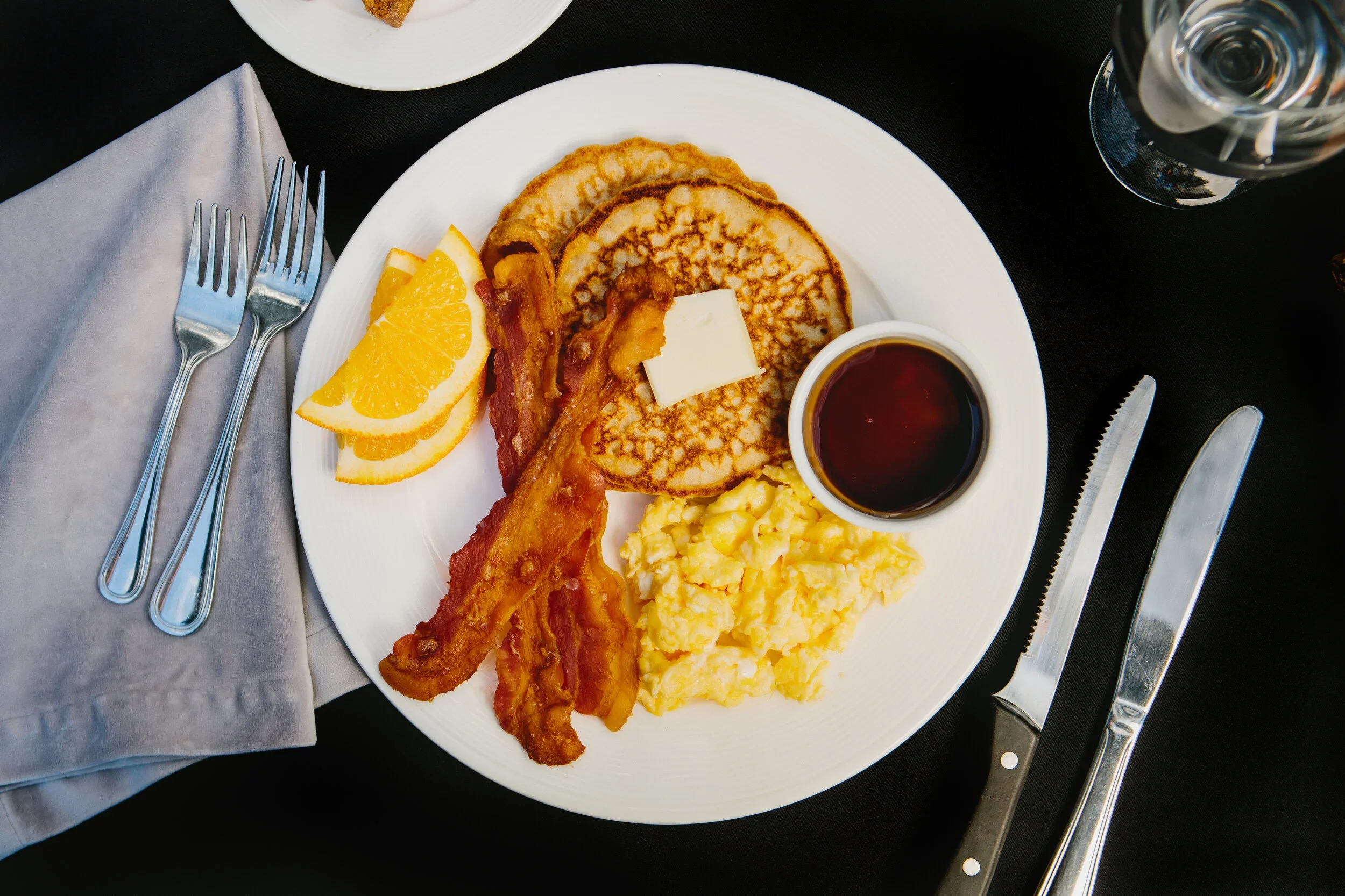 A breakfast plate with scrambled eggs, two slices of bacon, two pancakes with a pat of butter, orange slices, and syrup. Silverware is placed on a napkin to the left, and a glass of water is on the right.