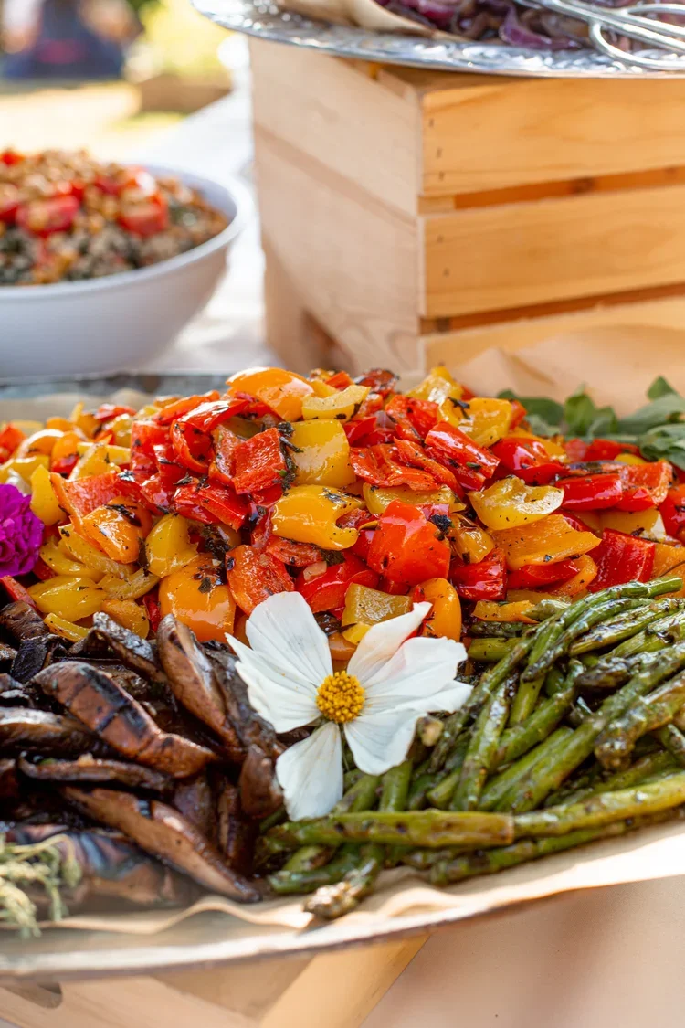 A tray of roasted vegetables including red and yellow bell peppers, grilled eggplant slices, and cooked asparagus, garnished with a white flower.