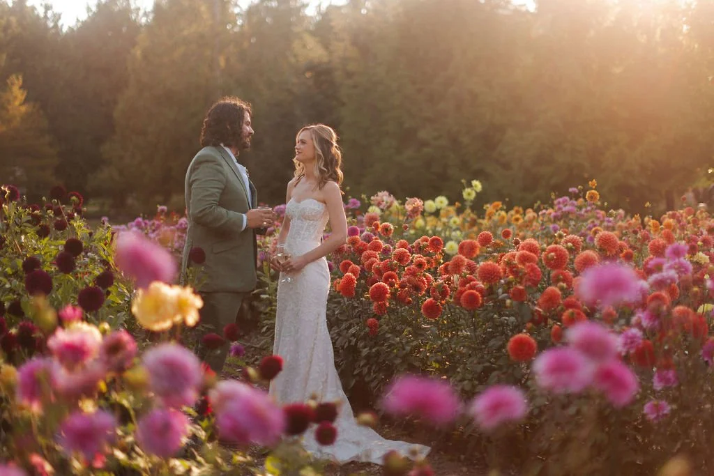 A bride and groom holding hands and smiling at each other in a flower garden at sunset.