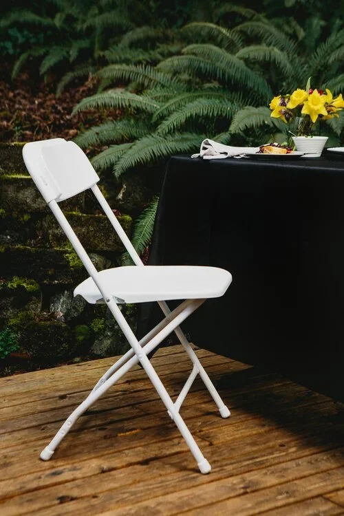 Empty white folding chair on wooden deck next to a table with a black tablecloth, yellow flowers, and dishes, with green fern plants in the background.