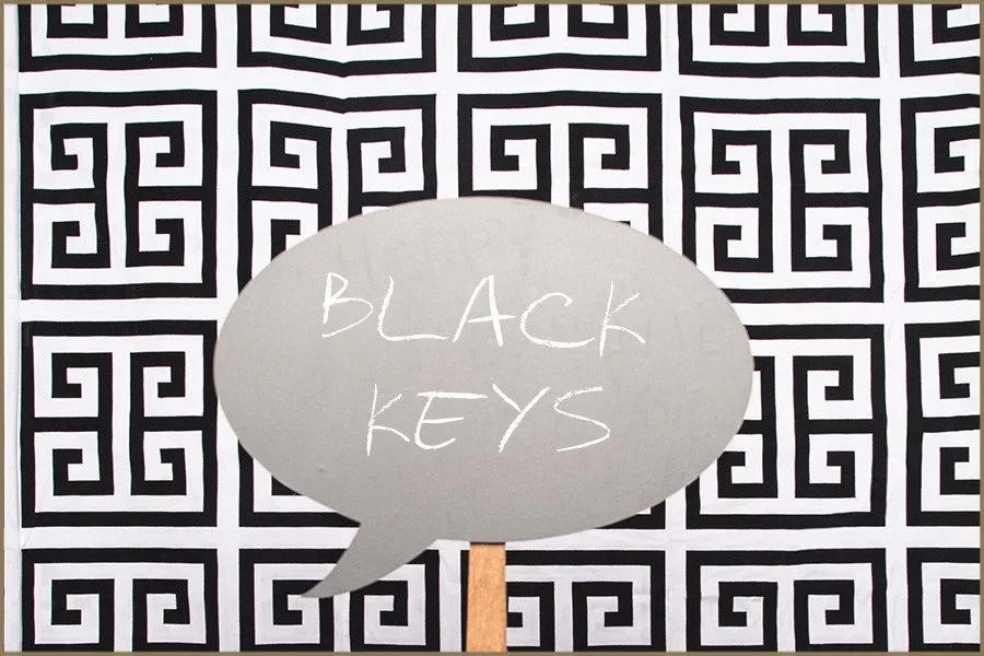 A speech bubble with the words "Black Keys" written inside, held up by a stick, against a black and white geometric patterned background.