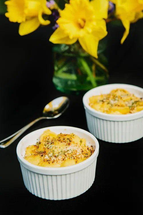 Two small white ramekins filled with baked macaroni and cheese, sprinkled with herbs, on a black surface next to a silver spoon, with a bouquet of yellow flowers in a glass vase in the background.