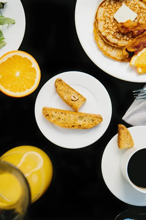A breakfast scene with pancakes topped with butter, orange slices, toast with nuts, a half orange, a glass of orange juice, and a cup of coffee on a black table.