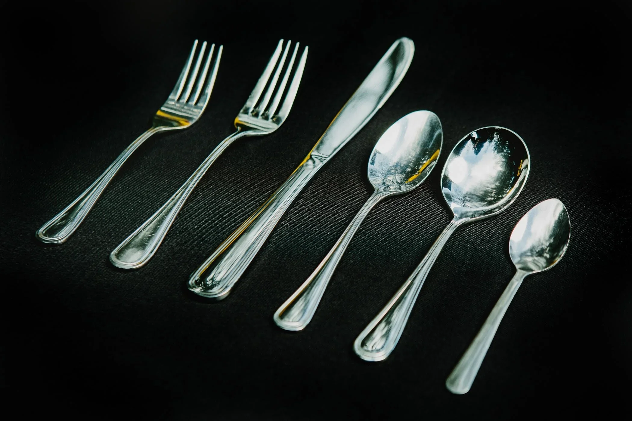 Six pieces of silverware arranged on a dark background: two forks, a knife, and three spoons.