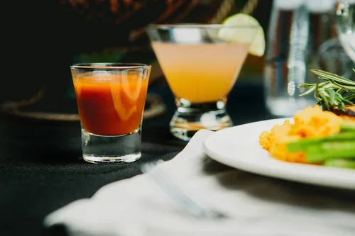Two cocktails, one small shot glass with layered red and orange drink, and a larger glass with a light yellow cocktail garnished with a lime wedge, on a black table with a white plate of food and green herbs nearby.
