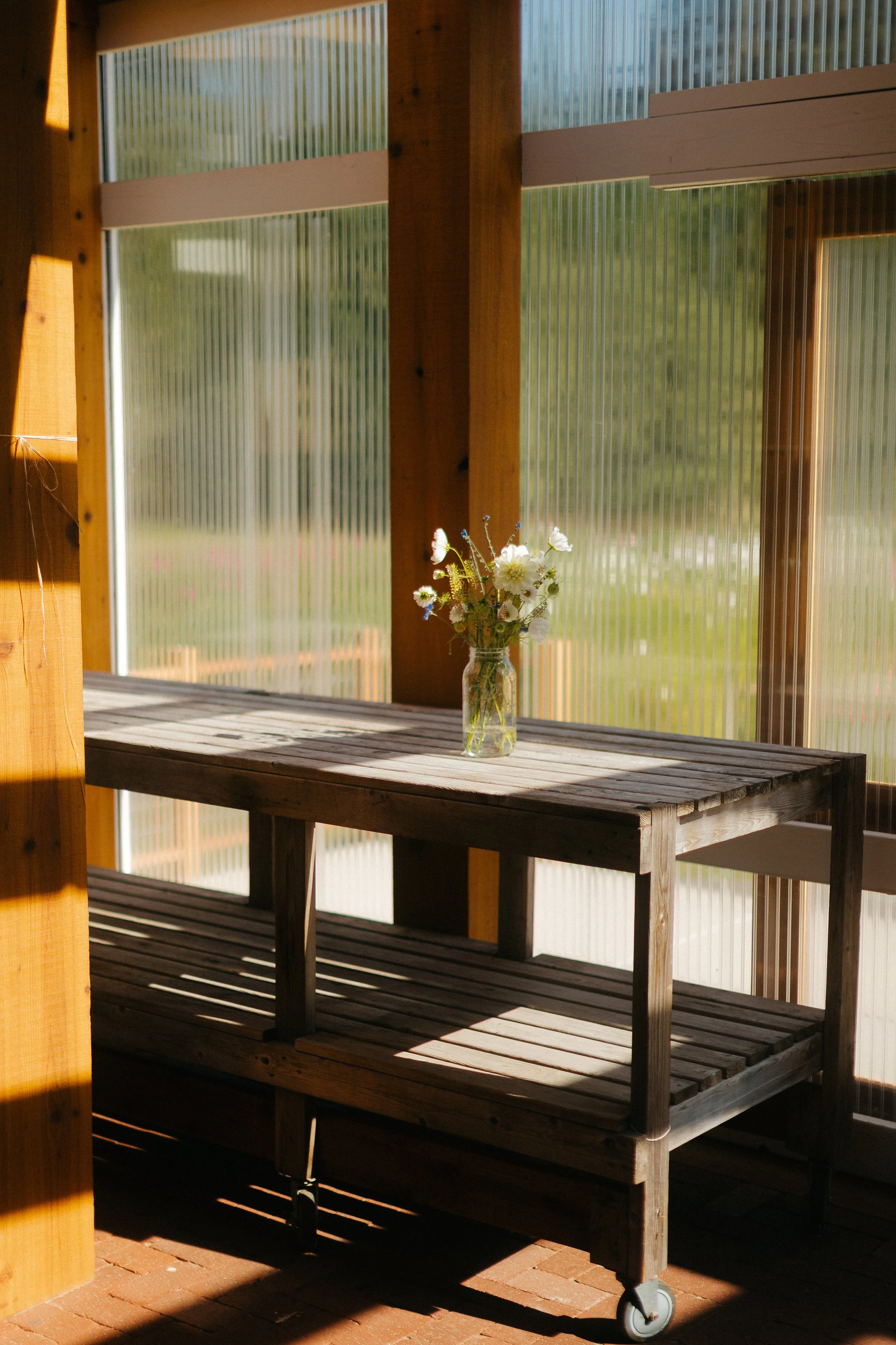 A glass jar holding a bouquet of white flowers on a wooden table inside a sunlit room with large windows.