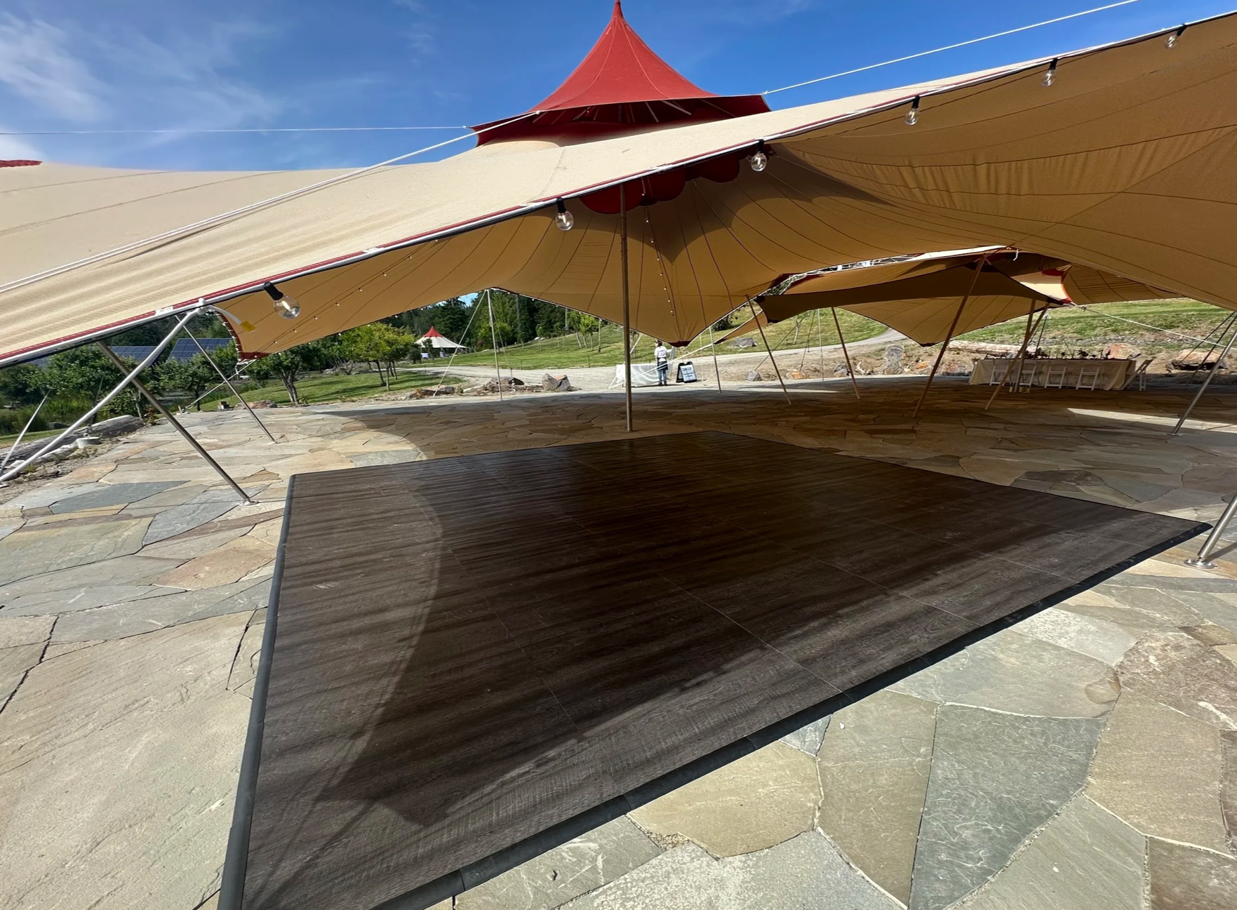 Large outdoor event tent with beige fabric and red accents, set up on a stone patio with sunlight and trees in the background.