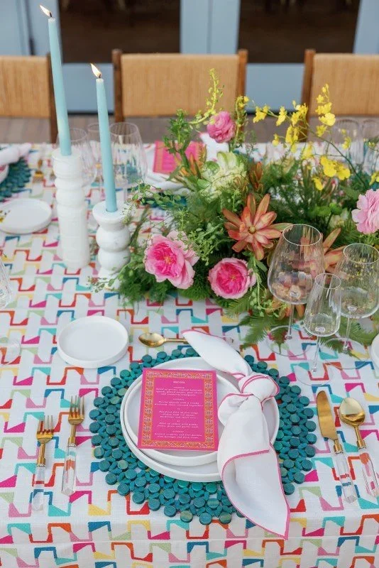 Colorful table setting with pink flowers, green foliage, tall blue candles, and glasses, featuring a pink menu on white dinnerware.
