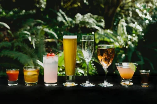 A variety of colorful cocktails and drinks on a black table outdoors with lush green foliage in the background.