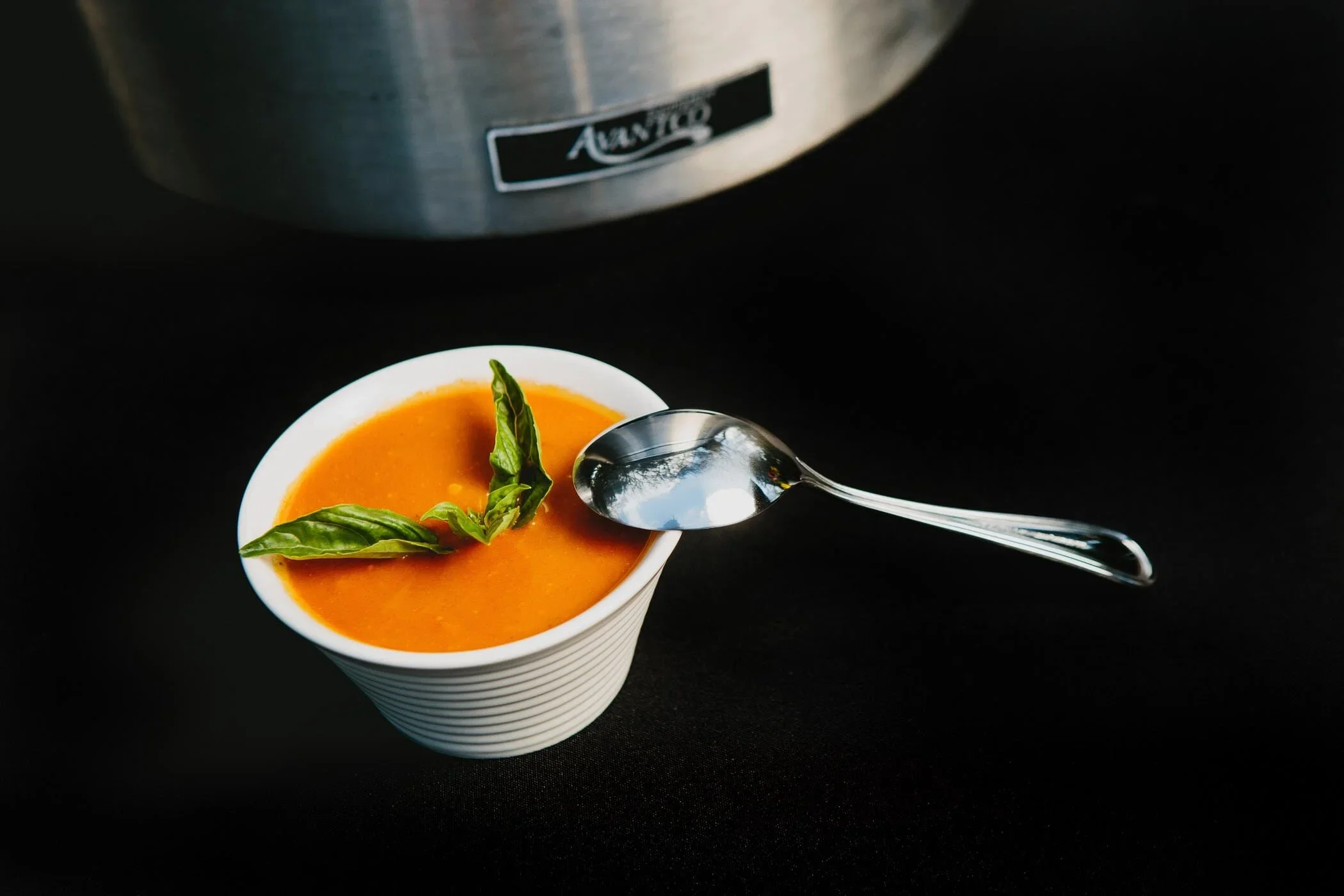 A cup of orange soup garnished with fresh basil leaves, with a silver spoon resting on the rim, placed on a black surface near a metal appliance.