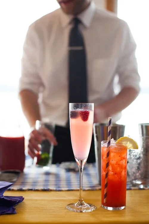 A bartender preparing colorful cocktails at a bar, with two drinks in the foreground: a pink cocktail in a champagne flute with a raspberry on top, and a red fruit drink with a lemon wedge and striped straw.