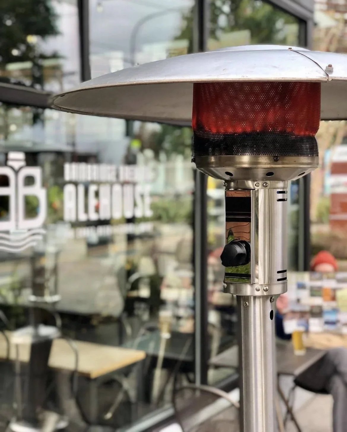 Outdoor patio heater with a metal heat reflector in front of a glass window, with blurred trees and sign reading 'The Broadway Alehouse' in the background.