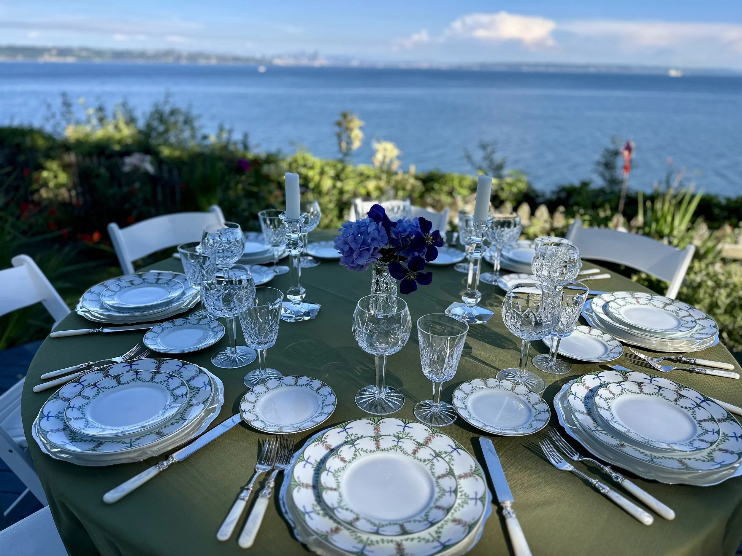 Dining table set outdoors with glassware, plates, cutlery, and floral centerpiece overlooking a body of water and distant skyline.