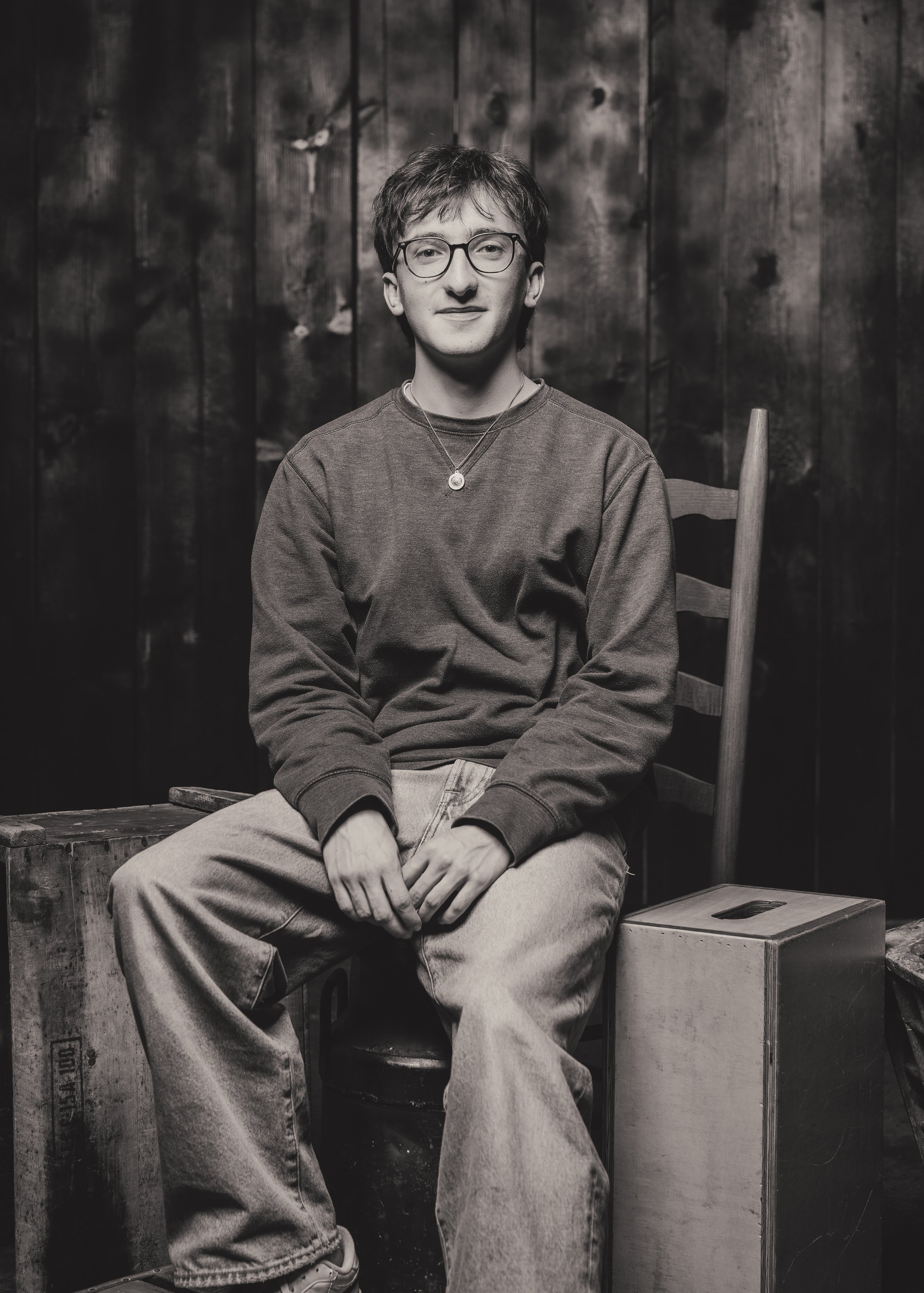 Black and white photo of a young man with glasses, sitting on a wooden chair with a relaxed pose, in front of a dark wooden background, wearing a sweatshirt and jeans.