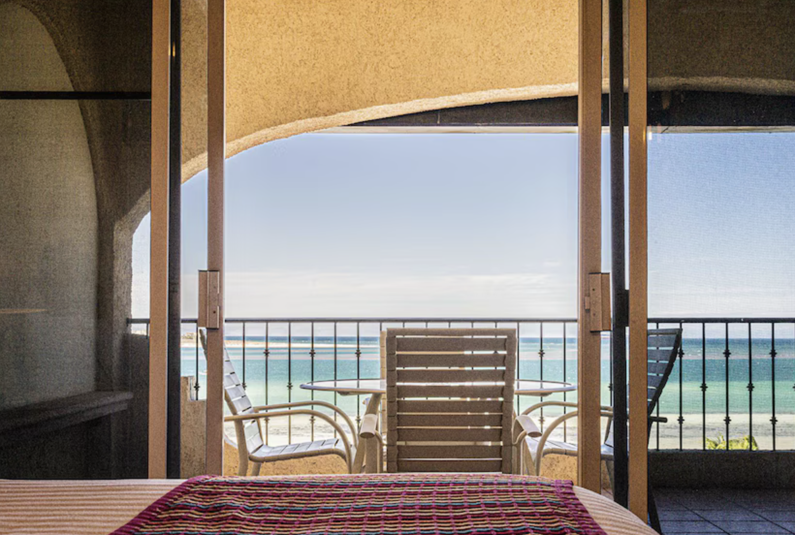 View from inside a room, looking out onto a balcony with ocean view, featuring two white chairs and a small table, with beach and ocean in the background.