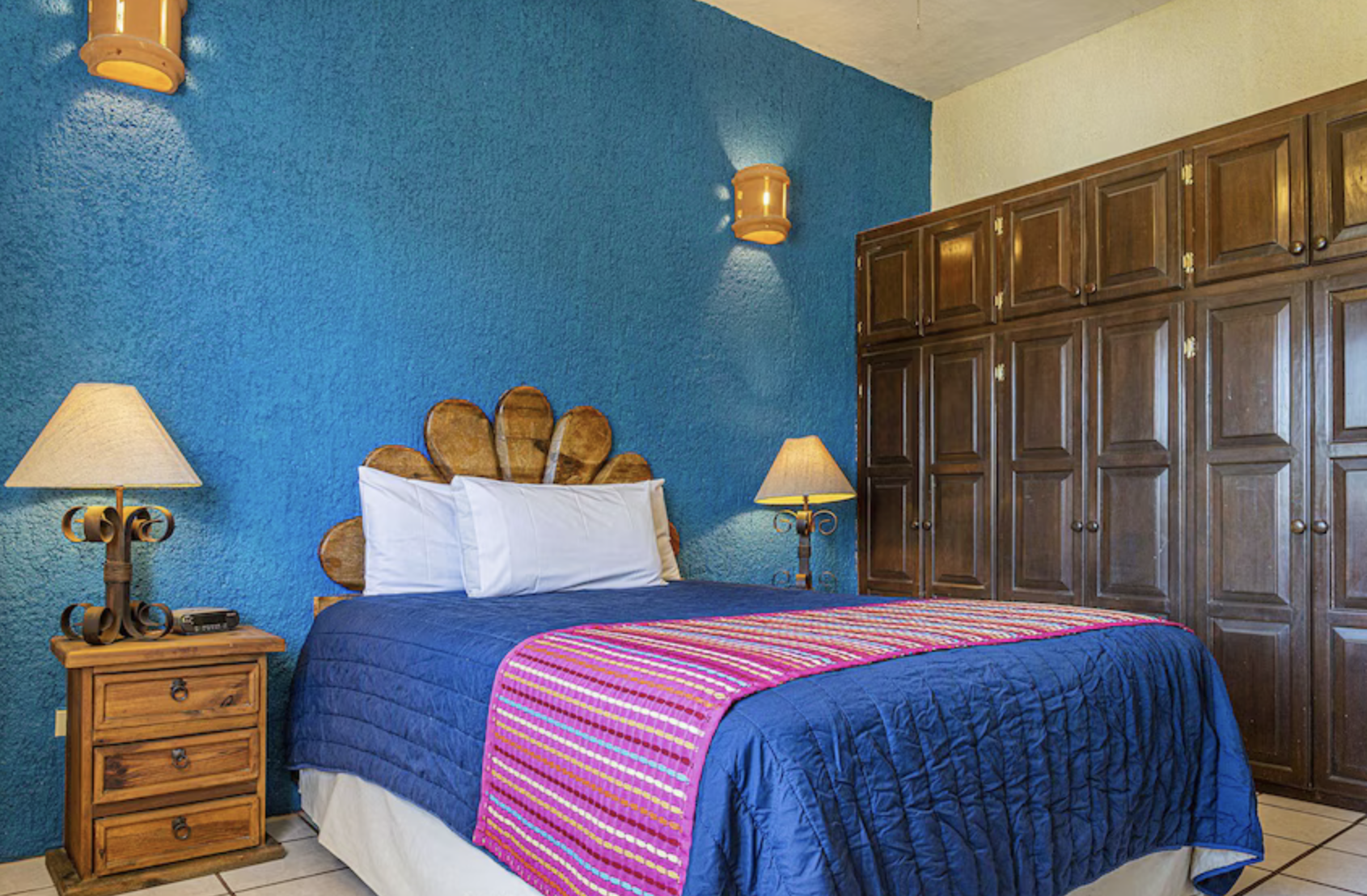Hotel bedroom with blue textured wall, wooden headboard, two wooden nightstands with lamps, large wardrobe, bed with blue quilt, and colorful striped blanket.