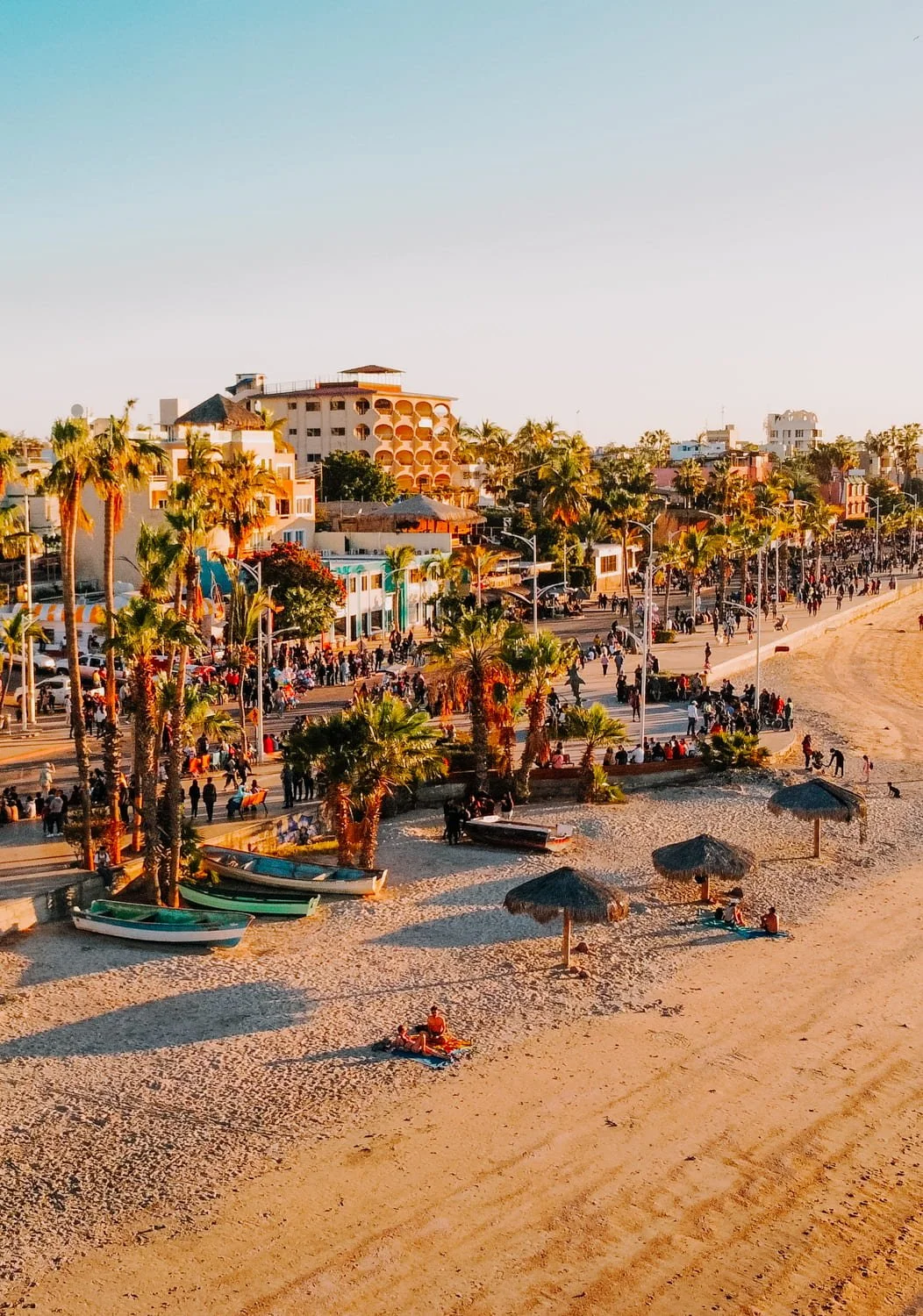 A lively beach scene during sunset with people walking along the boardwalk, palm trees, colorful buildings, and umbrellas on the sandy beach with boats