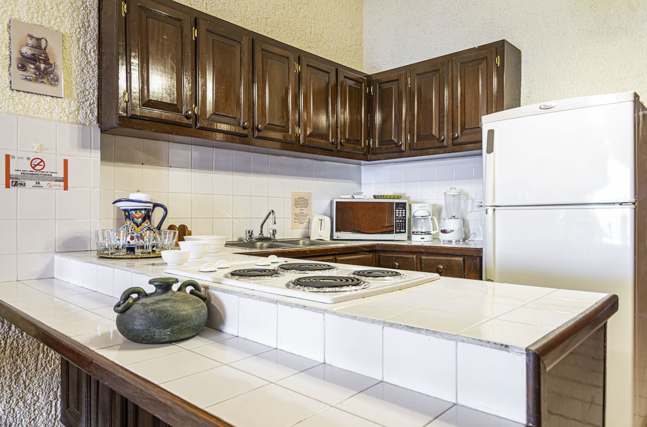 Kitchen with wooden cabinets, white tiled countertops, white appliances including refrigerator, microwave, blender, and coffee maker, with decorative pottery and glassware on the counter.