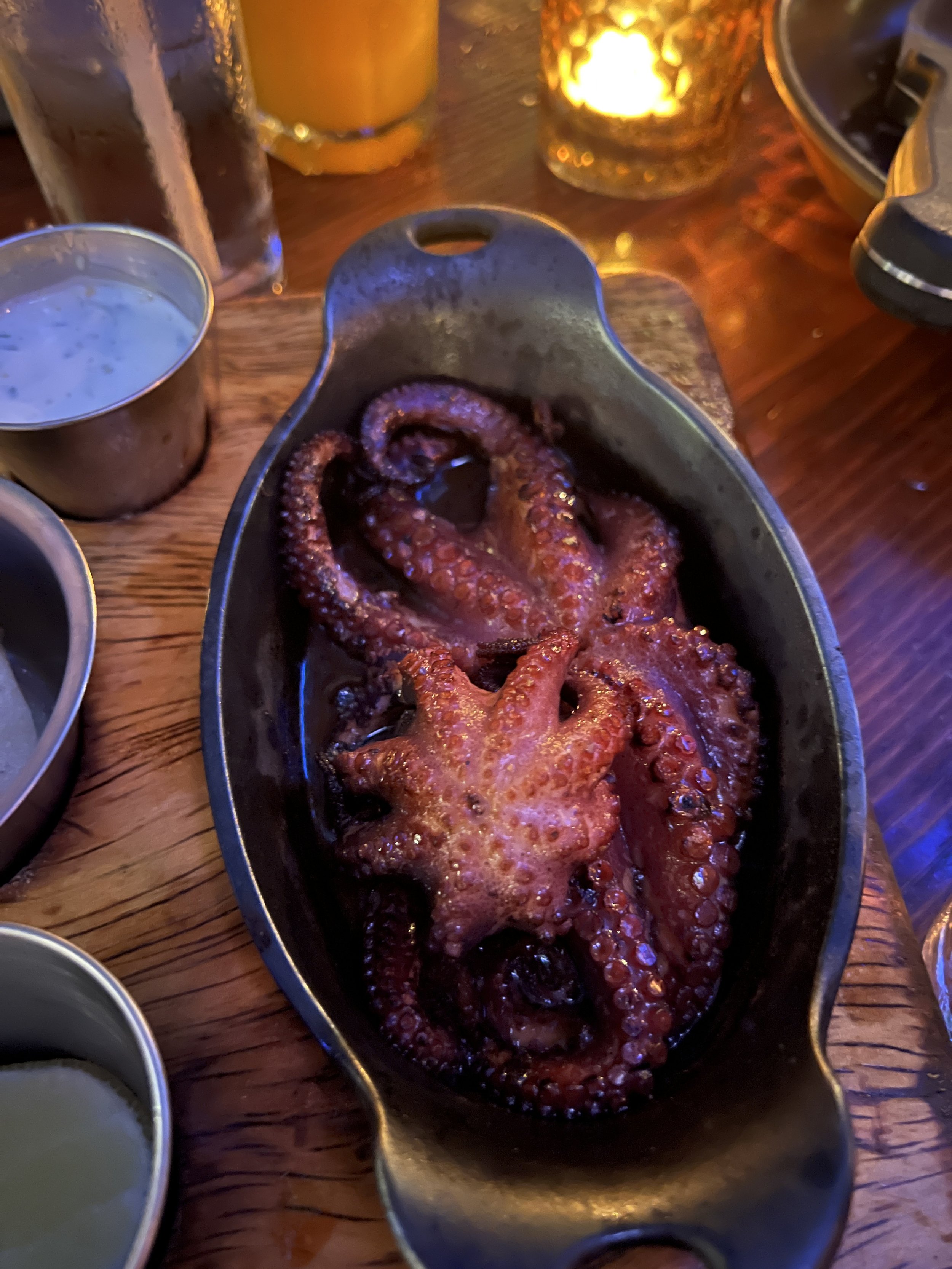 Cooked octopus tentacles served in a black dish on a wooden table, with drinks and condiments nearby.