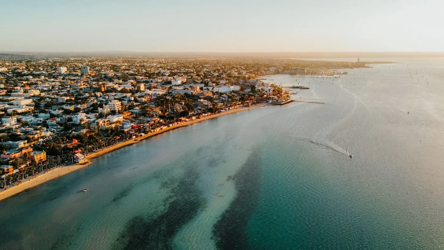 Aerial view of a city coastline with residential buildings, a beach, and a sailboat on the water during sunset.