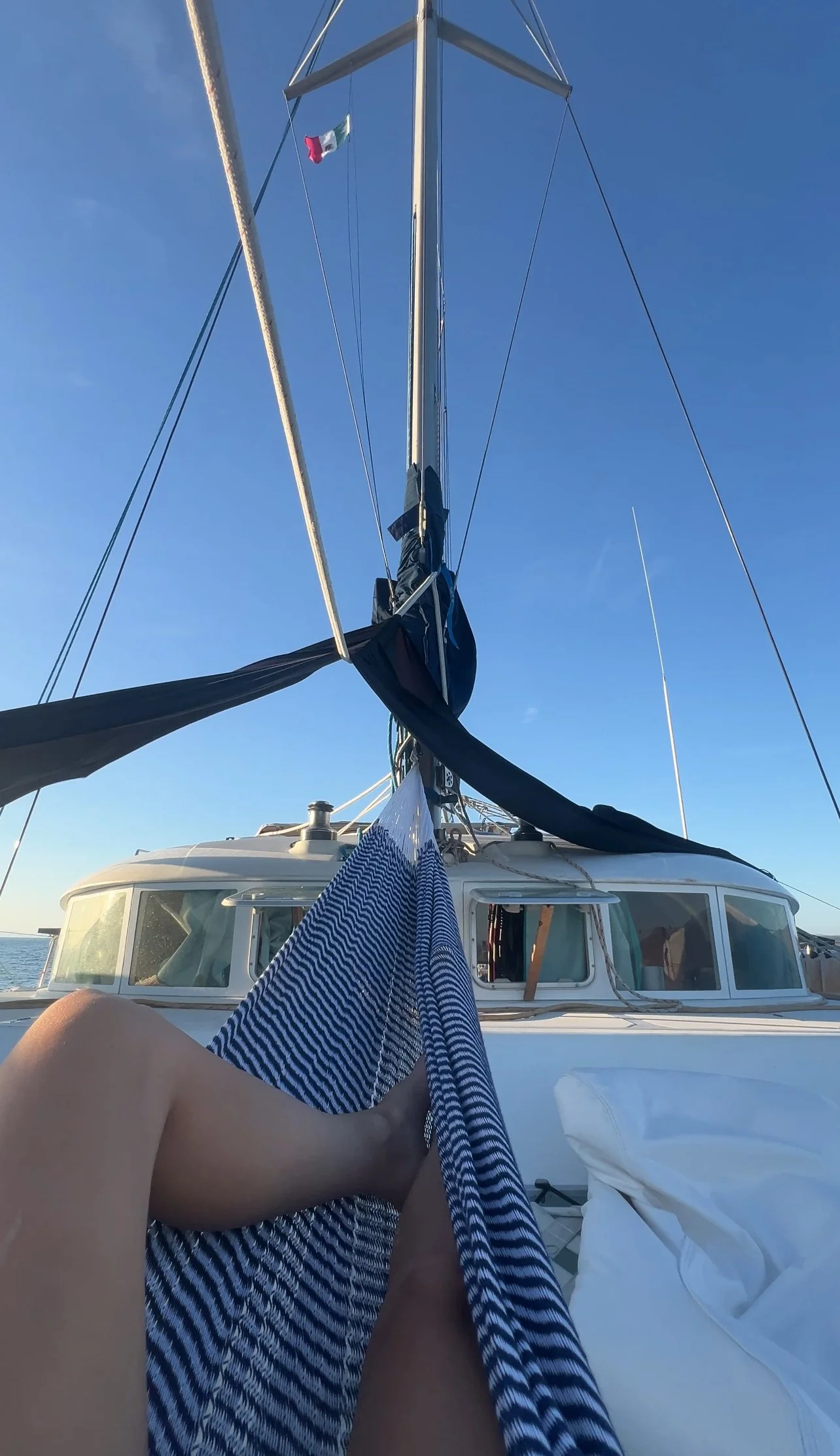 Person relaxing on a sailboat with legs stretched out and a blue and white striped hammock, the boat's mast and sails visible against a clear blue sky.