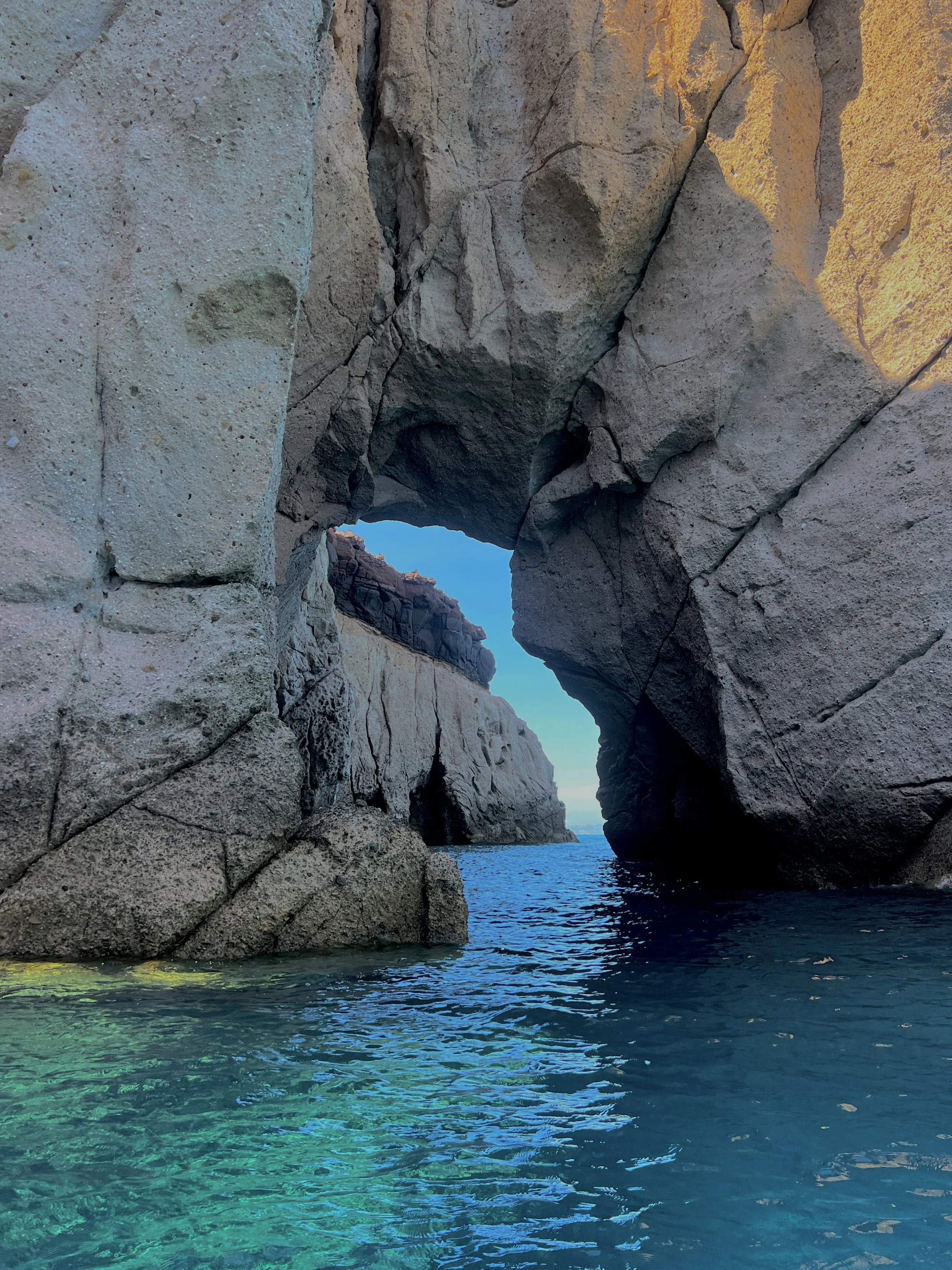 A natural sea arch formed by large rocky cliffs with turquoise water below and a clear blue sky in the background.