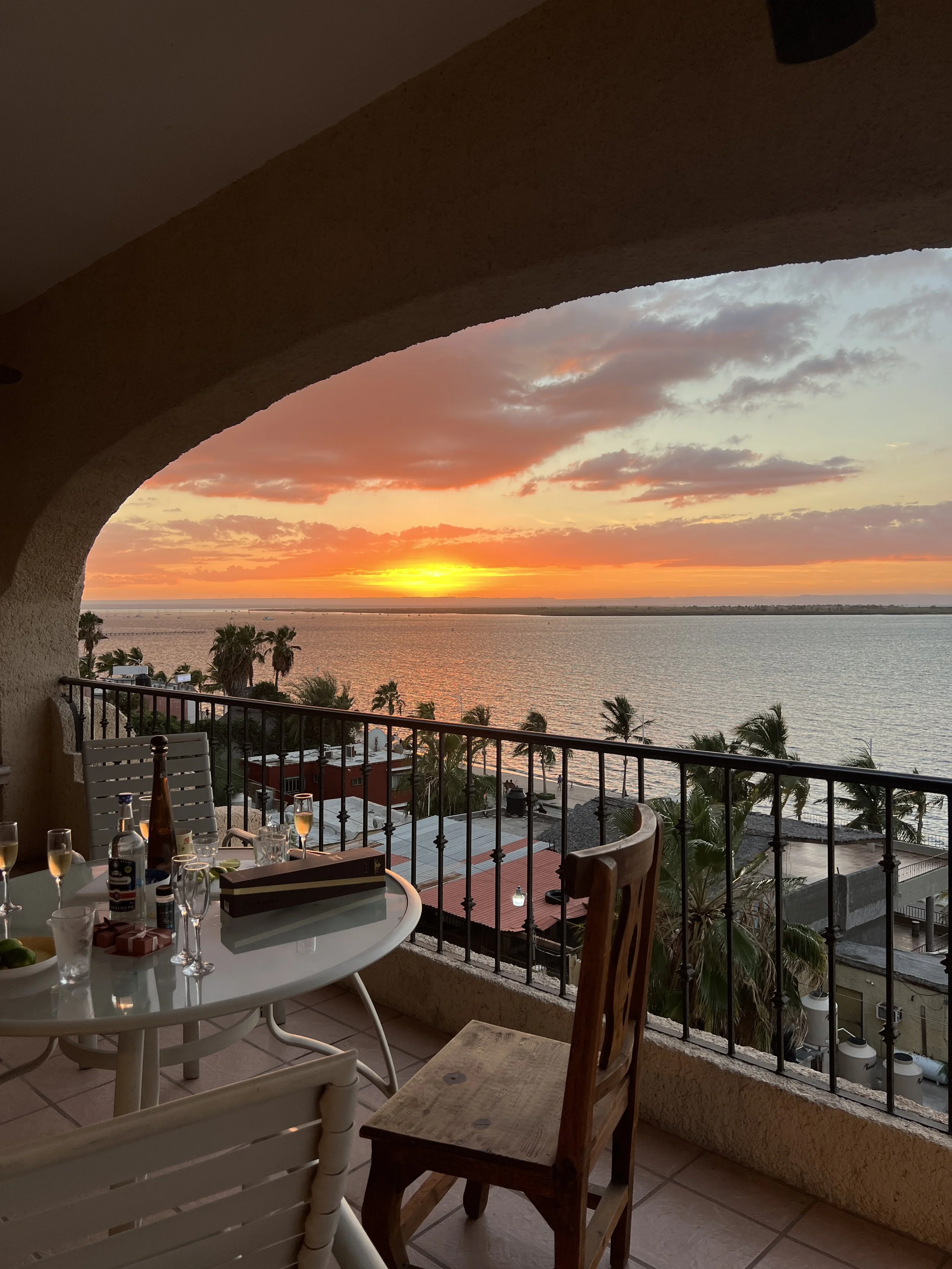 Balcony with a table and chairs overlooking a body of water at sunset, with palm trees and rooftops in the foreground.