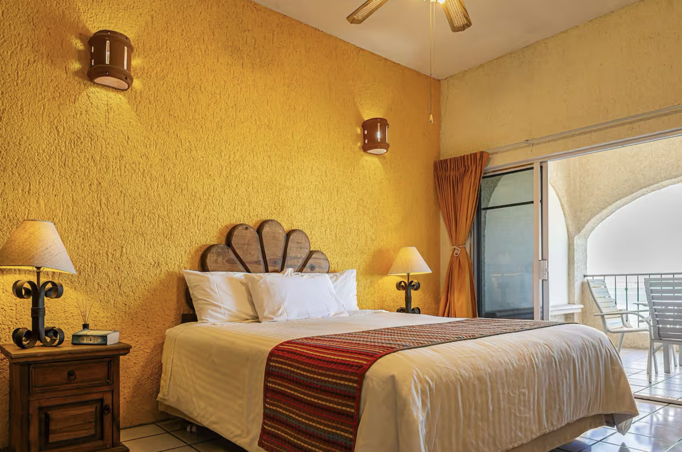 Hotel room with yellow textured walls, a large bed with white linens and colorful runner, wooden headboard, nightstands with lamps, and a sliding glass door leading to a balcony with patio furniture.