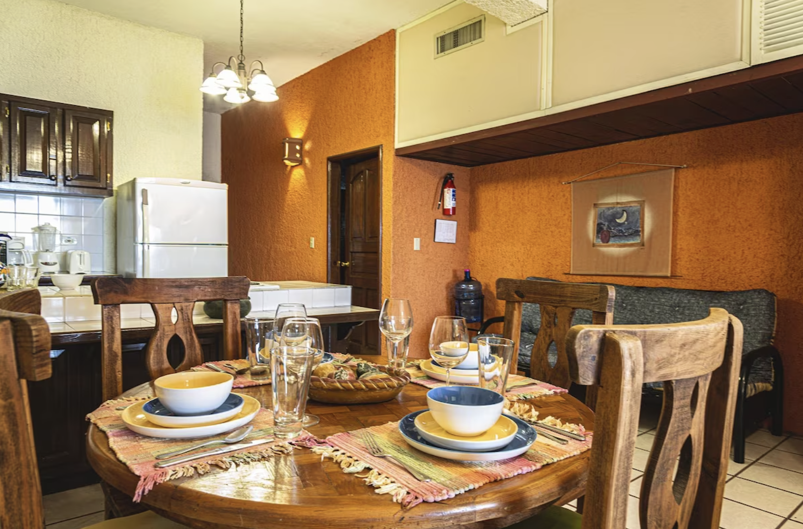 A dining table set with bowls, plates, glasses, and cutlery in a cozy kitchen with wooden chairs and orange walls.