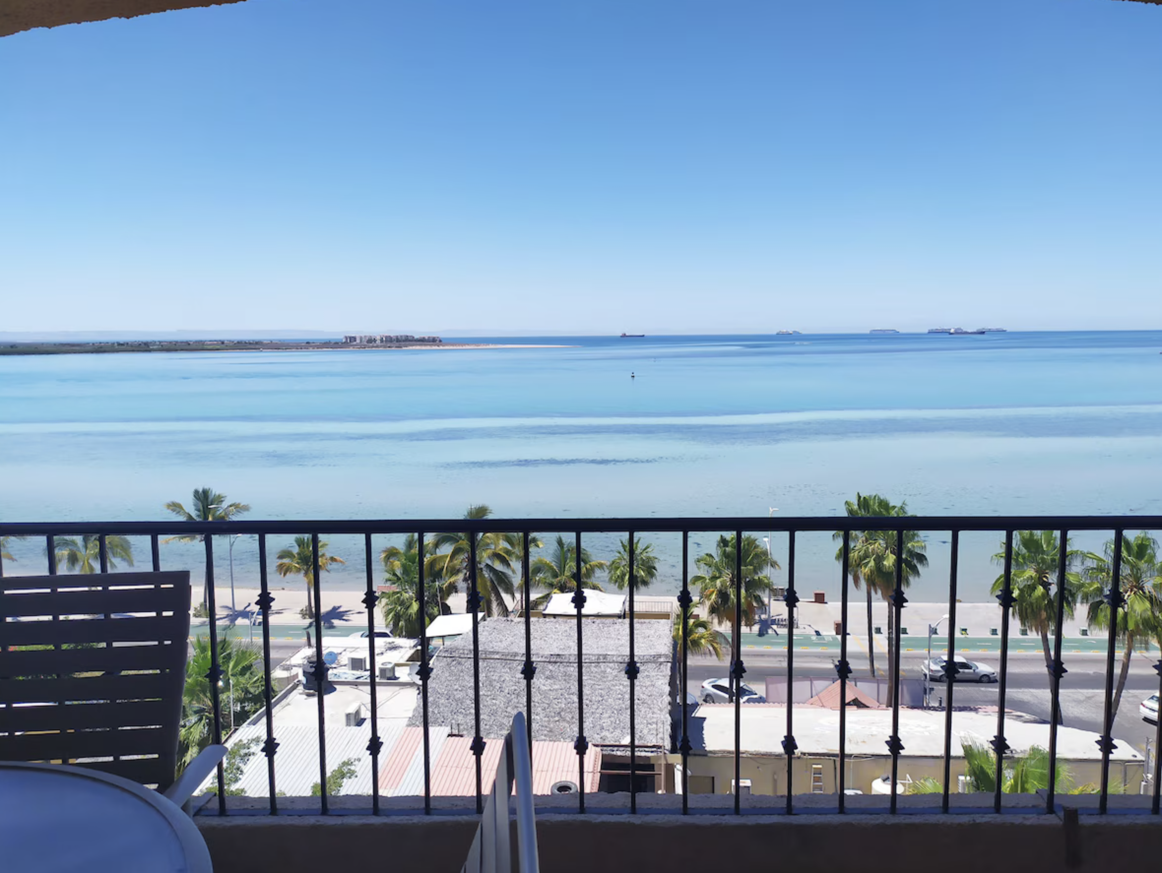 Balcony view overlooking a beach with palm trees, a street, and the ocean with ships in the background under a clear blue sky.