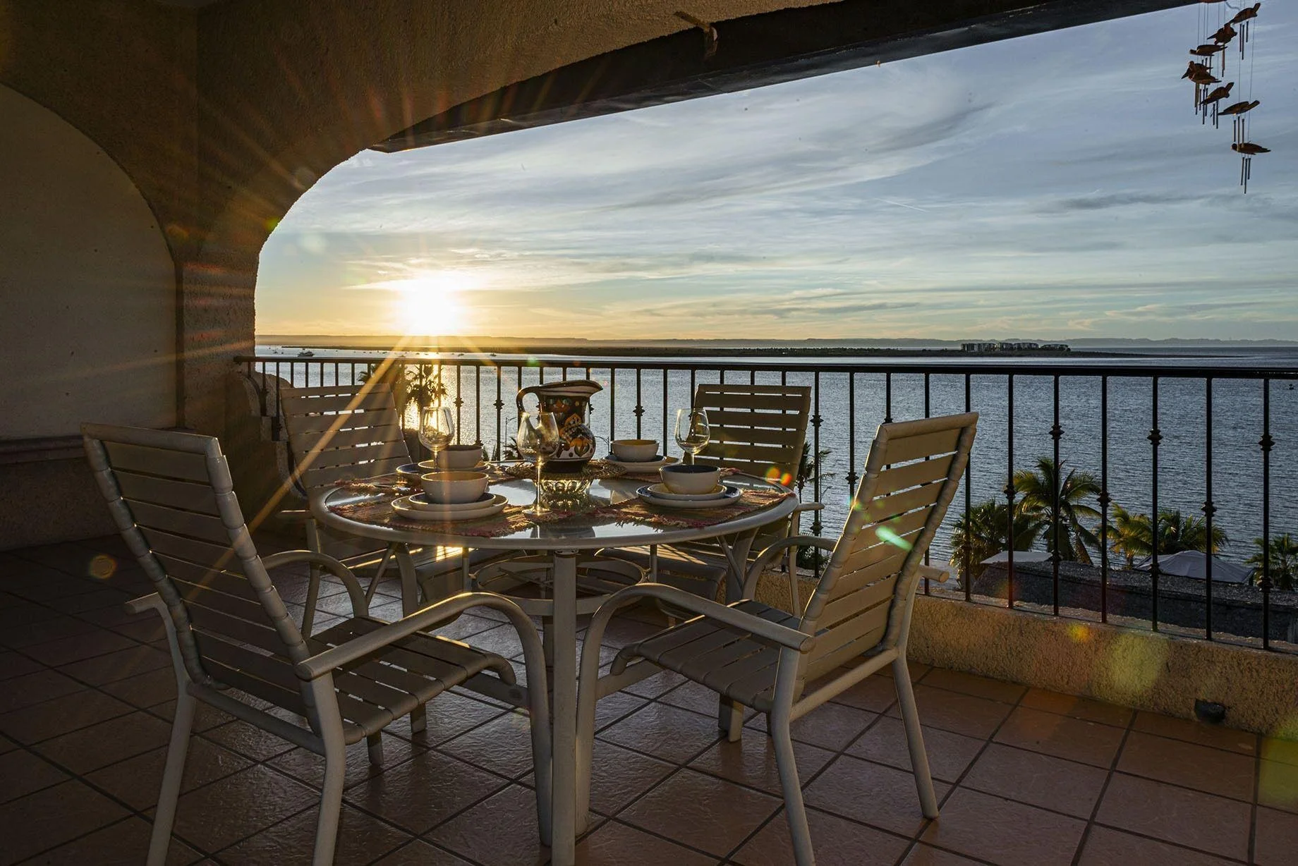 Dining table set for four on a balcony overlooking a body of water at sunset, with palm trees in the background.