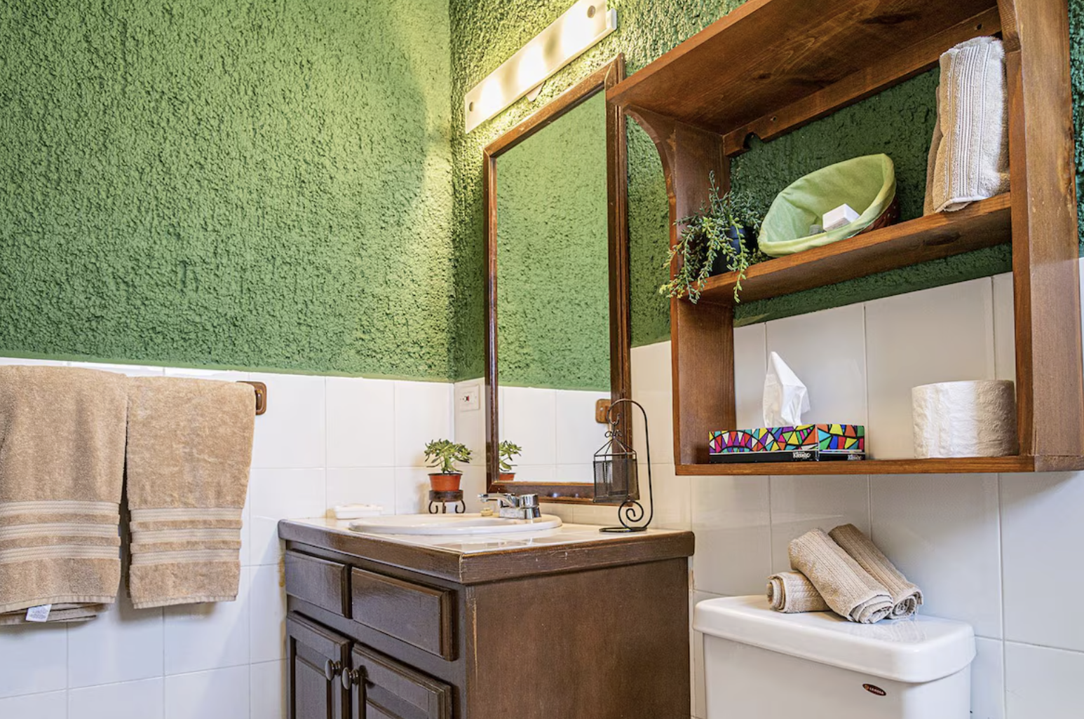 A bathroom with a green textured upper wall, white tile lower wall, and a wooden bathroom cabinet with a mirror. There are beige towels hanging on the wall, a small potted plant on the sink, and a wooden shelf above the toilet holding a green bowl, a rolled towel, decorative plant, tissue box, and toilet paper rolls.