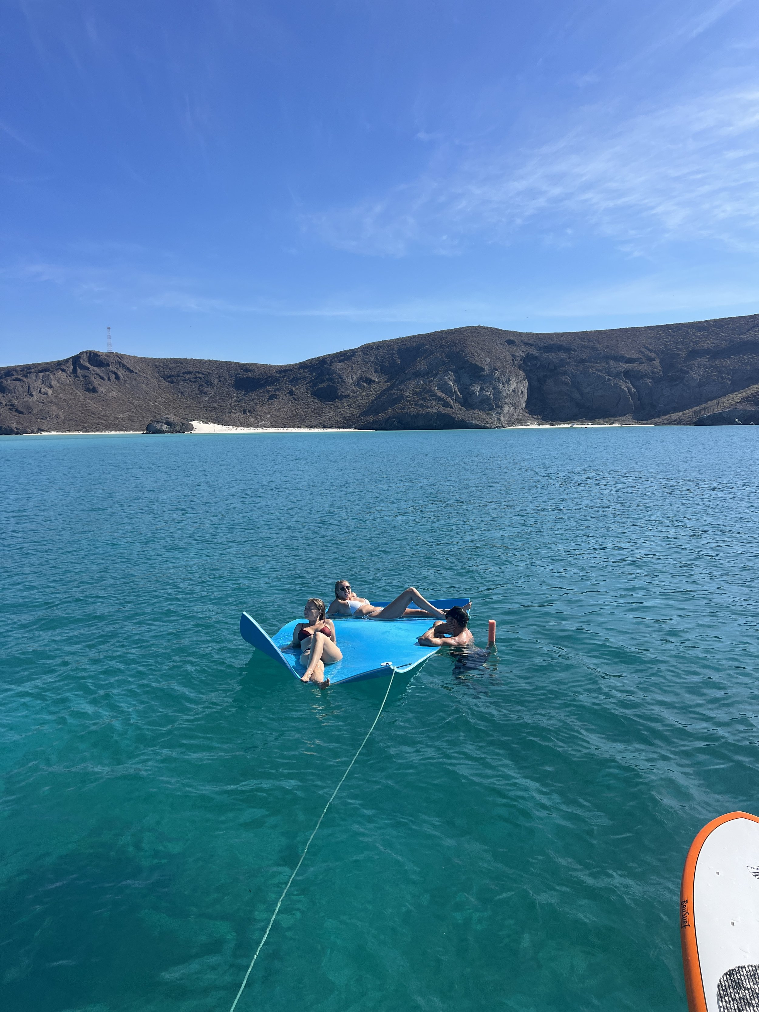 Three people relaxing on a small, upside-down blue boat in a calm, turquoise lake with a mountainous landscape and clear blue sky in the background.