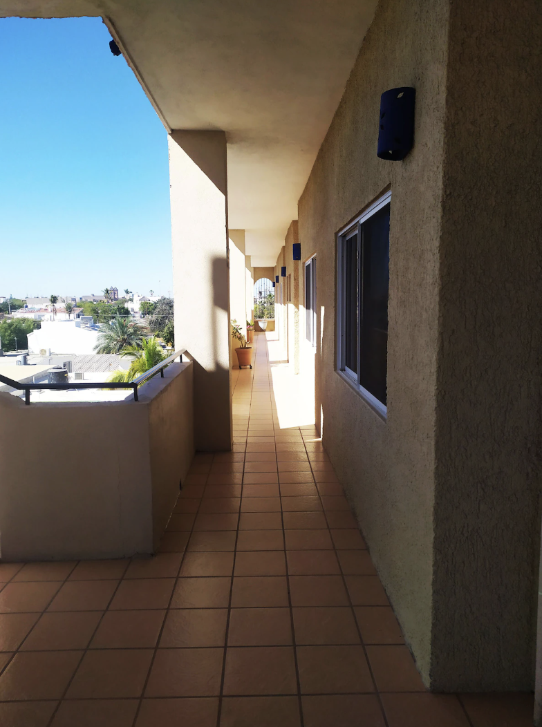 Balcony walkway with potted plants, windows, and exterior lighting fixtures overlooking a cityscape with palm trees.