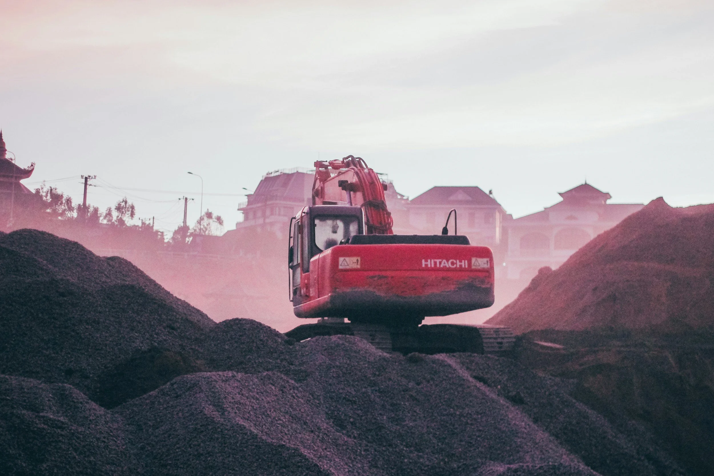 Red compact excavator on a mound of dirt or gravel at a construction site with houses in the background.